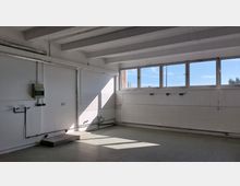 Leerer Raum mit weißen Wänden und Sichtbetondecke, großen Fenstern, die Tageslicht einlassen, sowie freiliegenden Rohren und Anschlüssen an den Wänden. Der Boden ist mit grauem Linoleum belegt, und ein schmaler Betonsockel verläuft an der Wand.