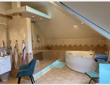 Modernes Badezimmer unter einer Dachschräge mit beiger Fliesenverkleidung. Zu sehen sind eine Eckbadewanne, eine offene ebenerdige Dusche, eine weiße Kommode, mehrere Handtuchhalter sowie ein Stuhl und ein klappbarer Hocker.