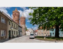 Straßenszene in einer historischen Altstadt mit zwei gut erhaltenen, runden Backsteintürmen im Hintergrund, umgeben von Fachwerkhäusern und einfachen Wohngebäuden. Im Vordergrund führt eine gepflasterte Straße an einem großen Baum und einem geparkten Auto vorbei.