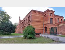 Backstein-Gebäude im neogotischen Stil mit Verzierungen aus helleren Backsteinen, großen Bogenfenstern und einem seitlichen Eingang. Im Vordergrund Grünflächen, ein kleiner Weg und ein Metalltor, umgeben von Sträuchern und Bäumen.