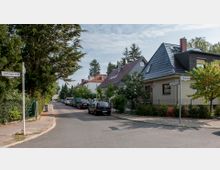 Ruhige Wohnstraße mit Einfamilienhäusern, von Bäumen und Sträuchern gesäumt. Geparkte Autos stehen entlang der Straße, und Straßenschilder zeigen die Namen „Föttingerzeile“ und „Zangenbergweg“.