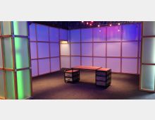 Ein moderner Raum mit hellen, halbtransparenten Wänden, die von einem Holzrahmen eingefasst sind, und farbiger Beleuchtung in Blau- und Grün-Tönen. Im Zentrum steht ein minimalistischer Schreibtisch mit Metallrahmen und Schubladen.