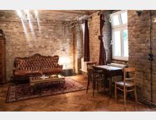 Wohnzimmer im Vintage-Stil mit Wänden aus unverputztem Backstein, einem braunen Ledersofa, einem verzierten Couchtisch auf einem orientalischen Teppich sowie einem Holztisch mit Stühlen. Die Fenster sind mit dunklen Vorhängen geschmückt, und die Decke zeigt freiliegende Rohre.