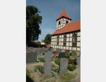 Ein Fachwerkkirchgebäude mit rotem Ziegeldach und Kirchturm, umgeben von einem Friedhof mit gepflegten Grabsteinen und Blumenbeeten. Im Hintergrund sind vereinzelte Bäume und ein blauer Himmel zu sehen.