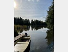 Ein ruhiger See, umgeben von Bäumen und Schilf, mit einem Holzboot am Ufer. Am Himmel ist die Sonne zu sehen, teils von leichten Wolken und Kondensstreifen umgeben.
