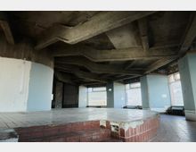 Industrieartiger, offener Raum mit unvollendeter Betonarchitektur und sichtbarer Rasterstruktur an der Decke. Der Boden besteht aus Fliesen, teilweise mit erhöhtem Podest, und die Wände haben große Fenster, durch die Tageslicht einfällt.