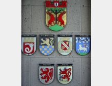 Wand mit sieben Wappen auf einer rechteckigen Steinfläche, darunter ein größeres Wappen oben mit roten Hirschen, stilisiertem Baum und Zinnen einer Stadtmauer. Darunter sechs kleinere Wappen verschiedener Städte und Regionen, jeweils mit individuellen Symbolen und Farben.