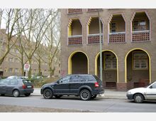 Wohnhaus im Stil der 1920er Jahre mit einzigartigen Fenstern und gelb umrahmten Arkaden; im Vordergrund parken mehrere Autos an einer Straße, flankiert von Bäumen mit kahlen Ästen. Ein 30er-Zonen-Verkehrsschild weist auf eine Verkehrsberuhigung hin.