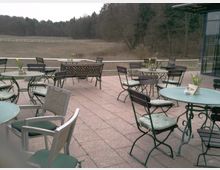 Terrasse eines Cafés oder Restaurants mit mehreren runden Tischen und Stühlen aus Metall, teilweise mit Polstern. Im Hintergrund ist eine offene Landschaft mit Wiese, Bäumen und einem Holzzaun zu sehen.