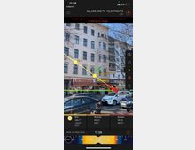 Ein Straßenabschnitt mit einem mehrstöckigen Wohnhaus im Hintergrund. Im Erdgeschoss befindet sich eine Bäckerei mit dem Schriftzug "Leckerback". Vor dem Gebäude sind geparkte Autos zu sehen, und Bäume ohne Laub ragen in den Himmel. Ein Overlay einer Sonnenstandsanwendung mit gelben und roten Markierungen sowie Uhrzeiten und Himmelsrichtungen liegt über dem Bild.