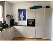 Moderne Küche mit weißen Einbauschränken und Arbeitsplatten in schwarzer Optik, integriertem Backofen und Herd. An der Wand hängt ein gerahmtes Poster mit einer Handzeichnung, daneben stehen Geräte wie eine Kaffeemaschine und ein Wasserkocher; links befindet sich ein Fenster mit Holzrahmen.