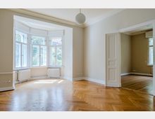 Heller, leerer Altbauwohnraum mit Fischgrätparkett, hohen Decken und großem Erker mit weißen Sprossenfenstern und Heizkörpern darunter. Rechts führt eine offene Kassettentür in ein angrenzendes Zimmer mit Holzboden und weiterem Fenster.