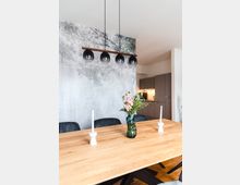 Esszimmer mit einem großen Holztisch, dekoriert mit einer Vase mit Blumen und zwei weißen Kerzenhaltern. Im Hintergrund eine Wand mit Marmoroptik, eine moderne Pendelleuchte mit kugelförmigen Lampenschirmen und eine offene Küche mit grauen Einbauschränken.