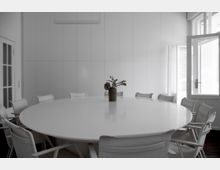 Runder Konferenztisch in einem hellen Raum mit weißen Wänden, umgeben von weißen Bürostühlen; in der Mitte des Tisches steht eine Vase mit Trockenblumen. Ein geöffnetes Fenster und eine Glastür lassen Tageslicht herein.