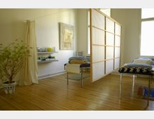 Ein heller, minimalistischer Raum mit Holzboden, ausgestattet mit Behandlungsliegen, weichen Decken und einer Trenntafel aus Holz und Stoff. Im Hintergrund sind ein Vorhang, ein Regal mit medizinischen Utensilien und ein Reliefbild an der Wand sichtbar.