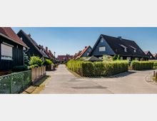 Wohnsiedlung mit freistehenden Einfamilienhäusern, die größtenteils schwarze Fassaden und rote Satteldächer haben. Der Fußweg verläuft mittig zwischen eingezäunten Vorgärten, die mit Hecken und Sträuchern bepflanzt sind.