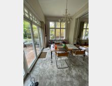 Esszimmer in einem hellen Raum mit großen Fenstern, die auf eine Terrasse blicken. Der Raum ist mit einem Holztisch, vier Stühlen mit Metallgestell, einem Teppichboden und einem Kronleuchter ausgestattet.