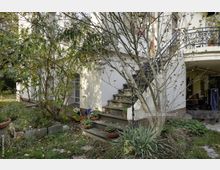 Wohnhaus mit weißer Fassade, teils von Bäumen und Sträuchern verdeckt. Sichtbar sind eine seitliche Steintreppe mit Blumentöpfen, ein schmiedeeisernes Geländer und ein überdachter Bereich mit Möbeln im Hintergrund.