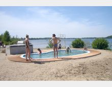 Ein rundes Schwimmbecken mit Holzumrandung steht auf einer sandigen Fläche in der Nähe eines Sees. Im Hintergrund sind Bäume, Büsche und der See mit blauem Himmel sichtbar, während Menschen im und um das Becken entspannen.
