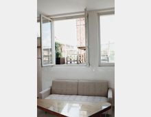 Wohnzimmer mit beigefarbener gepolsterter Couch und Glastisch im Vordergrund. Im Hintergrund befinden sich zwei geöffnete Fenster, die auf eine Terrasse mit Holzmöbeln und einer Hauswand aus Ziegelsteinen blicken.