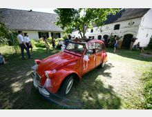 Ein roter Oldtimer (Citroën 2CV) dekoriert mit weißen Schleifen steht auf einer Wiese vor einem weißen Landhaus mit Giebeldach und angrenzenden Gebäuden. Im Hintergrund sind mehrere Personen in sommerlicher Kleidung, die sich in einer lockeren Gruppenatmosphäre unterhalten.