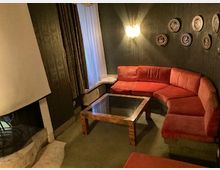 Wohnzimmer mit dunkler Wandgestaltung und einer halbrunden roten Polster-Sitzgruppe. Ein Glastisch steht zentral, an der Wand hängen dekorative Teller, und ein Kamin befindet sich links neben einem bodenlangen Fenster mit Vorhang.