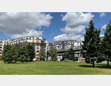 Wohnblöcke mit hellen Fassaden und bunten Fensterläden, flankiert von einem modernen Metallsteg, im Hintergrund. Im Vordergrund ein Park mit einer großen, grünen Rasenfläche, mehreren Bäumen und Menschen, die im Schatten sitzen.