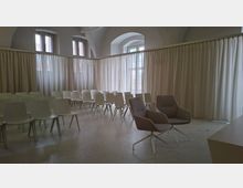 Heller Veranstaltungs- oder Seminarraum mit hohen Gewölbedecken und mehreren Fenstern, umgeben von langen weißen Vorhängen als Raumtrennung. In der Mitte stehen Reihen weißer Stühle sowie vorne zwei graue Sessel auf hellem, glattem Boden.