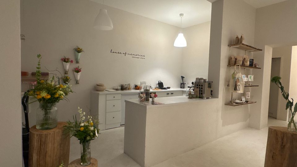 Heller, minimalistischer Ladeninnenraum mit weißem Tresen und Thekenbereich, Pendelleuchten und einer Kaffee- bzw. Kassenstation im Hintergrund. An den Wänden und auf Holzpodesten stehen Blumenarrangements, rechts hängen Regalbretter mit kleinen Produkten und Tüten.