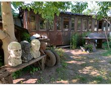 Ein rustikaler Garten mit einer alten, braunen Hütte im Hintergrund, mehreren Büsten aus Stein auf einer Holzbank, und Werkmaterialien wie Reifen und ein Metallgitter auf einem Arbeitstisch. Umgeben von Bäumen, Pflanzen und Schattenflächen.