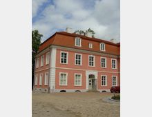 Ein zweistöckiges Gebäude mit rosa Fassade, weißen Fensterrahmen und einem roten Mansardendach. Vor dem Gebäude befindet sich ein gepflasterter Platz, und ein kleines rotes Auto steht rechts neben dem Eingang.