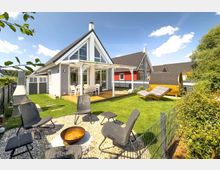 Ein modernes Ferienhaus mit spitzem Giebeldach und großer Glasfront, umgeben von einem gepflegten Garten. Im Garten befinden sich eine Terrasse mit Tisch und Stühlen, eine Feuerstelle mit Sitzgelegenheiten, eine Fasssauna und Liegestühle.