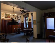 Gemütlicher Gastraum mit dunklen Holzmöbeln, Leuchtern an der Decke und dekorativen Bildern an den Wänden. Ein Fenster mit floralem Vorhang und Türen mit Glaseinsätzen sorgen für natürliches Licht.