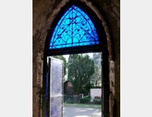 Ein gotisches Rundbogenfenster mit blauer, reich verziertem Glasmalerei und schwarzem Metallgitter öffnet sich zu einem Friedhof mit Grabsteinen und hohen Zypressen im Hintergrund.