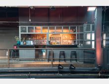 Eine kleine Bar in einem industriellen Raum mit einer Glasfront und Metallrahmen. Im Hintergrund sind beleuchtete Regale mit Flaschen und eine Kreidetafel mit Cocktailangeboten zu sehen, davor stehen Barhocker und eine Metallbrüstung.