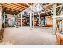 Innenraum einer alten Industrieanlage mit sichtbaren Rohren, Trichtern, Metallstützen und Lüftungssystemen. Der Raum hat eine Holzdecke, rote Treppen und eine weitgehend leere Bodenfläche, die von natürlichen Lichtquellen erhellt wird.