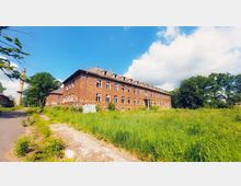 Großes, backsteinernes, verlassenes Gebäude mit mehreren Fenstern, teilweise beschädigt und mit Graffiti versehen, umgeben von einer überwucherten Wiese; im Hintergrund ein hoher Schornstein und Bäume unter einem blauen Himmel mit weißen Wolken.