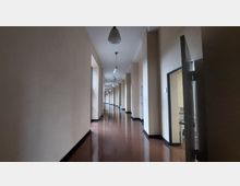 Ein langer, schmaler Flur mit beigen Wänden und glänzendem, braunem Boden. An der Decke hängen gleichmäßig verteilte kugelförmige Lampen, rechts sind Türen zu sehen, von denen eine geöffnet ist.