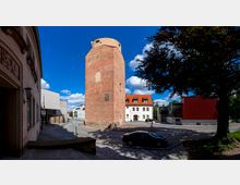 Backsteinturm inmitten einer städtischen Umgebung mit gepflasterten Straßen, links und rechts flankiert von modernen und historischen Gebäuden, darunter ein weißes Gebäude mit der Aufschrift „Kreismuseum“. Ein Baum wirft Schatten auf die Szenerie.