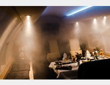 Innenraum eines luxuriösen Privatjets mit gedämpftem Licht und einer leichten Rauch- oder Nebelwirkung. Ein elegant gedeckter Tisch mit Gläsern, Geschirr und Blumenarrangements dominiert die Szenerie, während gepolsterte Sitze und Fenster die Raumstruktur bestimmen.