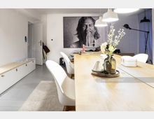 Heller Wohnraum mit Fokus auf einem großen Holztisch und weißen Stühlen, dekoriert mit einer Vase mit Trockenblumen. Im Hintergrund ein großformatiges Schwarz-Weiß-Porträt sowie eine weiße Tür und minimalistische Möbel.