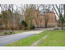 Ein Park mit einem gepflasterten Weg, vereinzelten Bänken und Bäumen ohne Laub. Im Hintergrund steht ein großes, historisches Backsteingebäude mit hohen Fenstern und verzierten Fassaden.