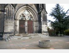 Vorderseite einer gotischen Kirche mit spitzbogigem Portal, zwei dunkelbraunen Holzflügeltüren mit dekorativen Verzierungen und Steinornamenten. Im Vordergrund ein gepflasterter Platz mit einer runden Betonkonstruktion und seitlich ein Baum sowie abgestellte Fahrräder.
