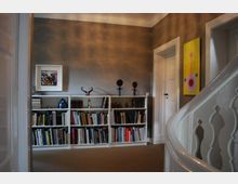 Ein gemütlicher Innenraum mit grauen Wänden und einer großen weißen Bücherwand, die mit Büchern und Dekorationsgegenständen wie Skulpturen und Bildern gefüllt ist. Im Vordergrund ist ein weißes, geschwungenes Treppengeländer zu sehen, und an der Wand hängen zwei moderne Kunstwerke.