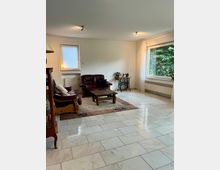 Helles Wohnzimmer mit großformatigem, hellgrauem Fliesenboden, zwei dunklen Ledersofas und einem niedrigen Holztisch auf einem Teppich. Große Fenster mit weißen Vorhängen und Heizkörpern darunter sowie ein kleiner Beistelltisch mit Nähmaschinenuntergestell stehen an den Wänden.