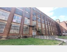 Backstein-Industriegebäude mit großen, säuberlich angeordneten Sprossenfenstern und einer Außentreppe, die zu einer Metalltür im zweiten Stock führt. Vor dem Gebäude erstreckt sich eine Rasenfläche, und der Himmel ist leicht bewölkt.