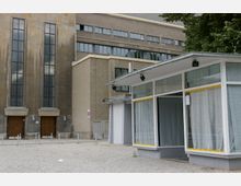 Außenansicht eines städtischen Gebäudekomplexes mit einem historischen Teil aus braunem Stein und großen Fenstern sowie einem moderneren Anbau mit großen Glasfassaden. Im Vordergrund ist ein leerer gepflasterter Platz zu sehen.