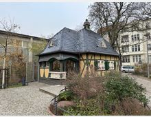 Kleines Fachwerkhaus mit geschwungenem Schieferdach, grünen Fensterläden und Zierpflanzen vor den Fenstern, gelegen an einem gepflasterten Gehweg mit umliegender Begrünung. Im Hintergrund befinden sich ein moderner Gebäudekomplex sowie Altbauten und kahle Bäume.