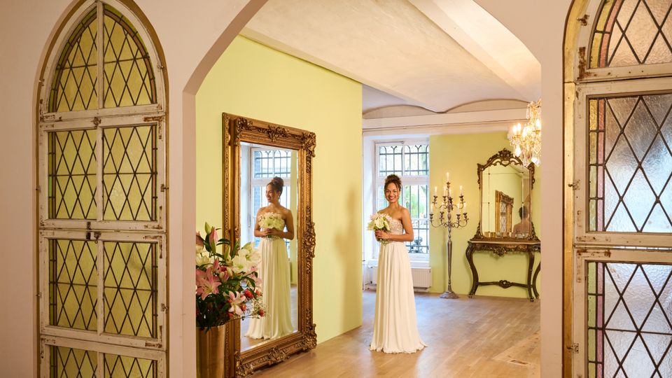 Eleganter Innenraum mit hohen Rundbögen, hellgrünen Wänden und Holzboden, flankiert von großen gotischen Türen mit Rautenglas. Im Raum stehen verzierte Goldspiegel, ein Konsolentisch, ein Kerzenleuchter und ein Kristalllüster; im Hintergrund sind große Fenster mit Sprossen zu sehen.