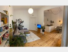 Wohnzimmer mit Holzdielenboden und heller Einrichtung, bestehend aus einem grauen Ecksofa, einem Regal mit Büchern und Pflanzen, sowie einem Fernseher. Im Hintergrund ist eine offene Küche mit weißen Schränken, einer Arbeitsplatte und einem Esstisch zu sehen; an der Wand hängen Hängelampen mit sichtbaren Kabeln.